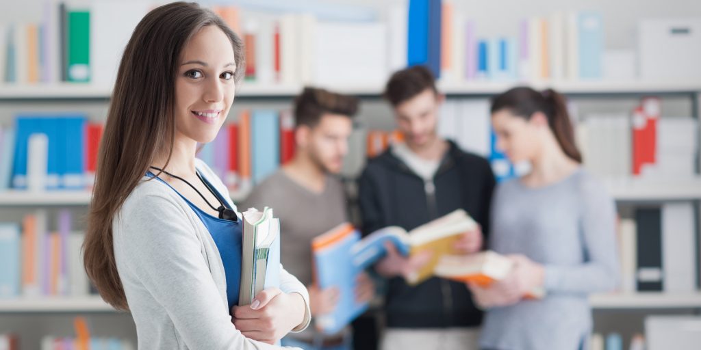 Confident smiling student girl posing in the library, holding books and looking at camera, learning and education concept